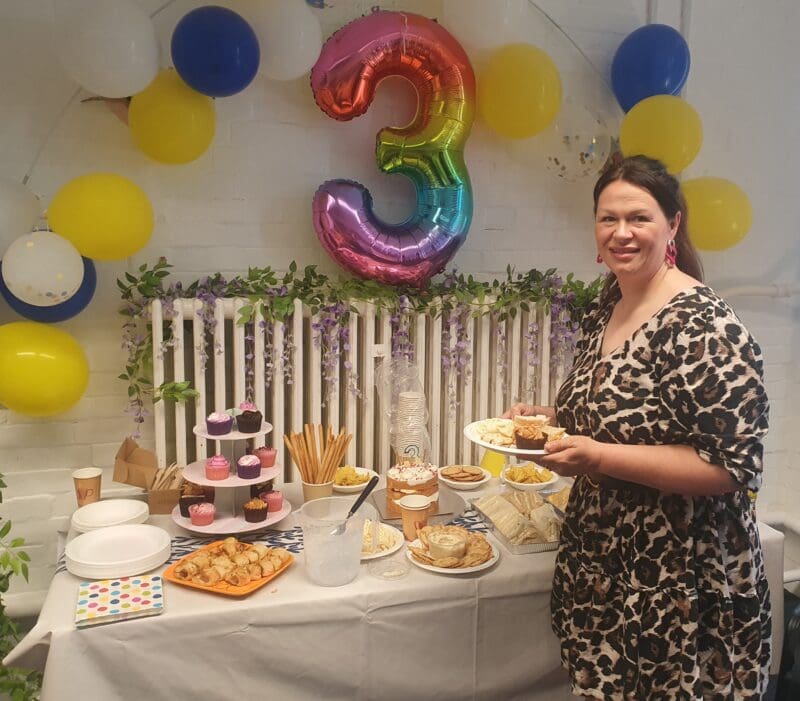Woman smiling next to table with cake. Above her is a balloon shaped as the number three.